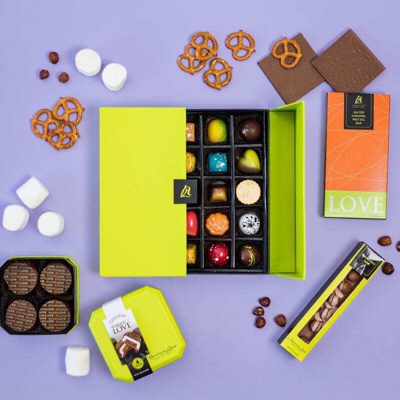 Overhead view of half open lime green signature truffle box with Hazelnut Bar, S'mores Love and Salted Caramel Pretzel Inclusion Bar pictured. Ingredients such as pretzels, marshmallows and hazelnuts surround chocolate box on a purple background. 